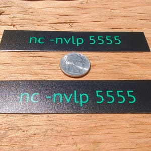 以下が含まれることがあります： 緑色の「nc-nvlp 5555」の文字が入った、2つの黒い長方形の金属板。硬貨がプレートの間にあります。プレートはキラキラした質感で、木製の表面に置かれています。
