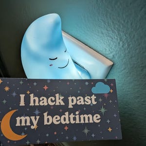 May include: A blue crescent moon night light with a smiling face and closed eyes plugged into a wall outlet. In front of the light is a rectangular sign with a dark blue background, colorful stars, a yellow crescent moon, and the text I hack past my bedtime.