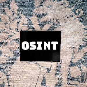 May include: A rectangular black sticker featuring the white text OSINT in a bold, sans-serif font. The sticker is placed against a textured fabric background that displays a repeating pattern of a mythical creature in shades of blue and beige.