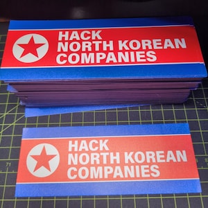 May include: Rectangular stickers with a red and blue design, stacked on top of each other. The stickers display a white star in a circle and the text "HACK NORTH KOREAN COMPANIES" in white.