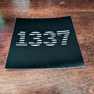 May include: A square black sticker featuring the number 1337 in a stylised white font. The digits are composed of horizontal lines, creating a striped effect across the surface of the sticker. The design is centred on a solid black background.