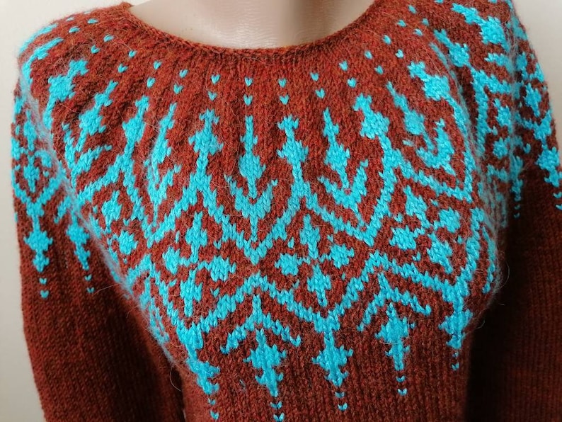 Pu&ograve; includere: Un maglione marrone con un motivo geometrico turchese sul petto e sulle spalle. Il maglione ha uno scollo tondo e maniche lunghe. Il design turchese &egrave; un motivo ripetuto di frecce e diamanti stilizzati.