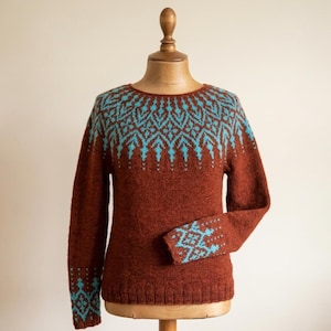Pu&ograve; includere: Un maglione marrone con un motivo geometrico turchese sullo sprone e sui polsini. Il maglione &egrave; esposto su un manichino di legno. Il design presenta un motivo ripetuto di forme a diamante e freccia.