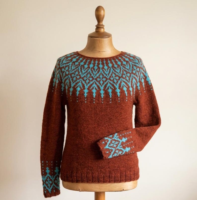 Pu&ograve; includere: Un maglione marrone con un motivo geometrico turchese sullo sprone e sui polsini. Il maglione &egrave; esposto su un manichino di legno. Il design presenta un motivo ripetuto di forme a diamante e freccia.