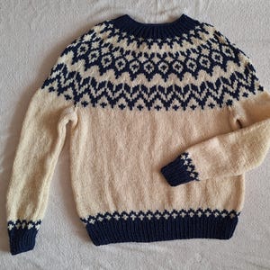 Peut inclure: Un pull en tricot crème avec un motif géométrique bleu marine autour du cou et de l'ourlet.