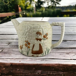 May include: A white ceramic jug with a stone-like texture and a brown and white illustration of a girl holding flowers. The jug has a handle and a spout.