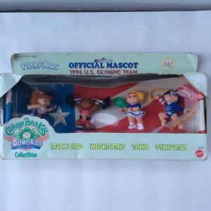 May include: A set of four Cabbage Patch Kids OlympiKids figurines in a clear plastic box. The figurines are dressed in red, white, and blue outfits and are holding various sports equipment, including a weight, a tennis racket, and a ribbon. The box has a red, white, and blue American flag design and the text "Official Mascot 1996 U.S. Olympic Team".