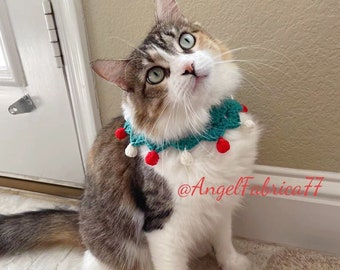 May include: A tri-colored cat wearing a teal crocheted collar with red pom-poms. The collar has a white crocheted snowflake design. The cat is looking up and has green eyes. The image includes the text "@AngelFabrica77".