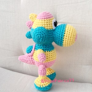 May include: A crocheted dinosaur toy with a pink, yellow, and blue color scheme. The dinosaur has a long neck and tail, and its eyes are black with white pupils.