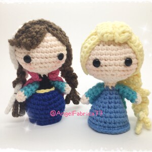 May include: Two crocheted dolls, one with brown hair and a blue dress, the other with blonde hair and a blue dress. Both dolls have large eyes and are wearing dresses with intricate details.
