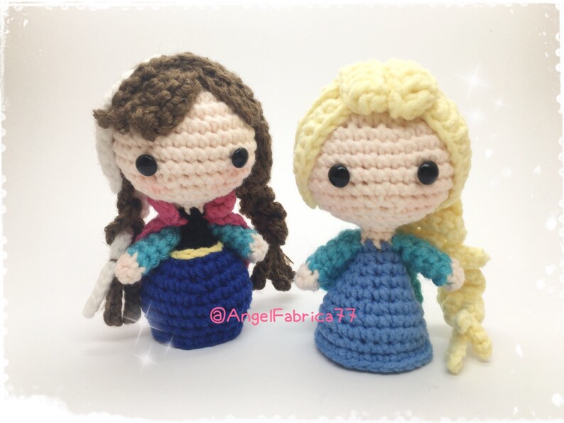 May include: Two crocheted dolls, one with brown hair and a blue dress, the other with blonde hair and a blue dress. Both dolls have large eyes and are wearing dresses with intricate details.