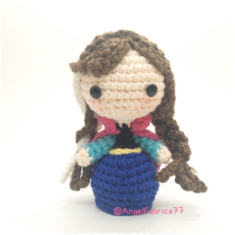 May include: Crocheted doll with brown hair, a pink and blue dress, and black eyes. The doll is wearing a blue skirt and a yellow belt. The doll is standing on a white background. @AngelFabrica77