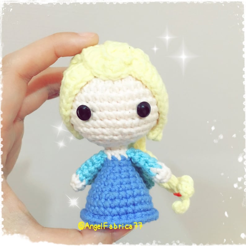 May include: A crocheted doll with blonde hair, blue and white dress, and black eyes. The doll is holding a yellow braid in its right hand.