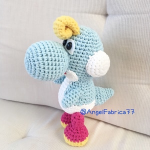May include: A crocheted blue and white Yoshi plush toy with yellow horns and pink shoes. The toy is standing on a white surface.