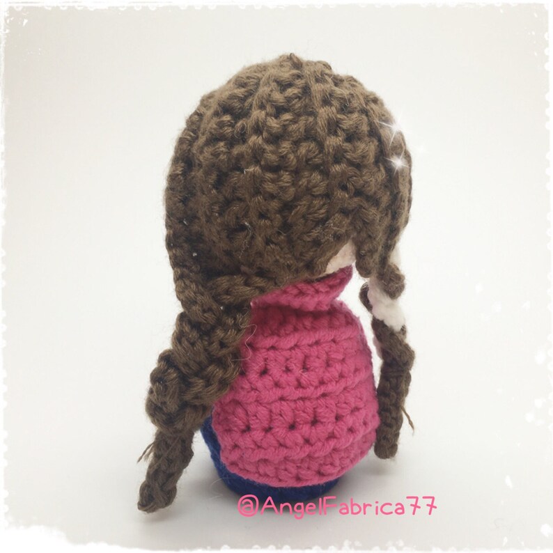 May include: A crocheted doll with brown hair in braids, wearing a pink sweater and blue jeans. The doll is facing away from the camera.
