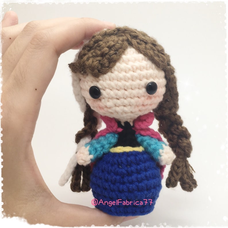 May include: Crocheted doll with brown hair, wearing a blue dress with a pink and blue jacket. The doll has black button eyes and is holding a white crocheted object.