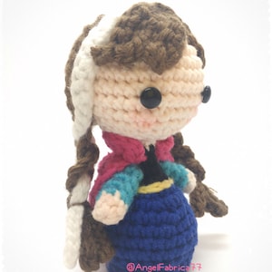 May include: Crocheted doll with brown hair, a white braid, and a blue dress. The doll is wearing a pink and blue jacket and has black button eyes. The doll is standing on a white surface. The text "@AngelFabrica77" is visible on the bottom of the image.