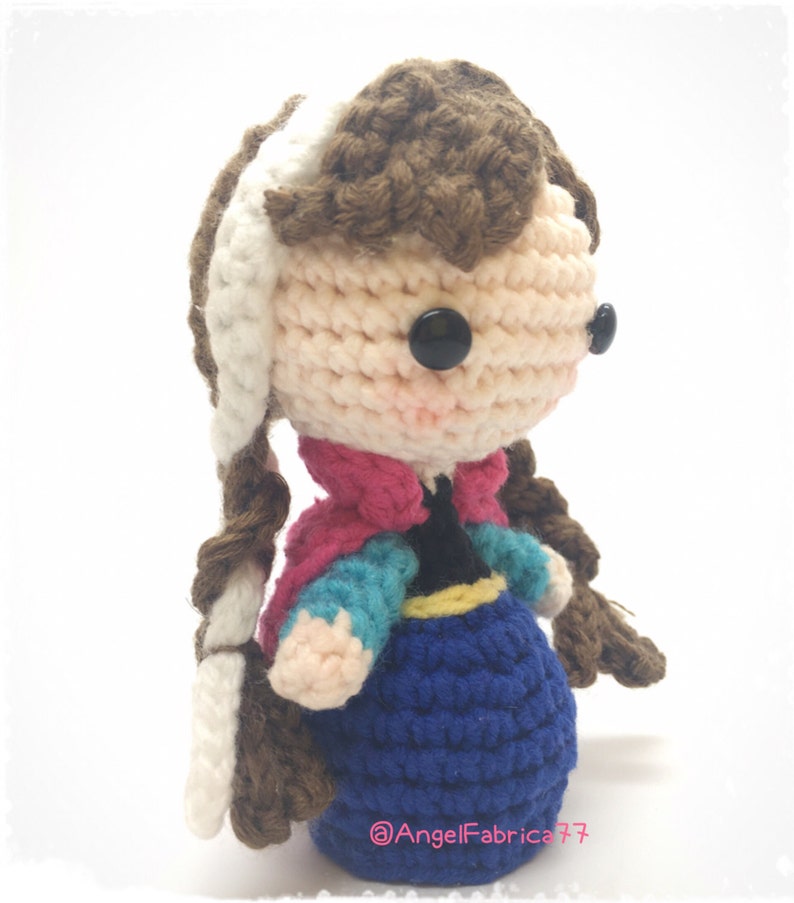 May include: Crocheted doll with brown hair, a white braid, and a blue dress. The doll is wearing a pink and blue jacket and has black button eyes. The doll is standing on a white surface. The text "@AngelFabrica77" is visible on the bottom of the image.