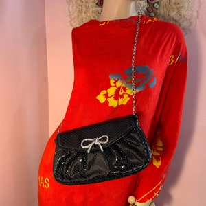 Op de afbeelding: Een zwarte clutch van imitatieleer met een strik van strass en een zilveren ketting.