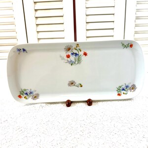 May include: A rectangular white ceramic serving platter with rounded corners. The platter is decorated with small floral bouquets in shades of blue, red, yellow, and white. The platter is displayed on a white lace tablecloth.