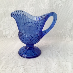 May include: A cobalt blue glass creamer with a diamond pattern and a raised design on the front. The creamer has a handle and a pedestal base.