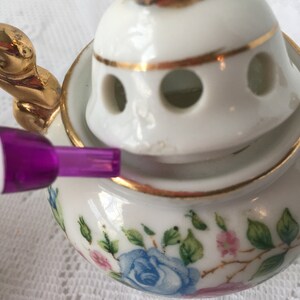 May include: A white porcelain incense burner with a gold trim and floral design. The lid has a gold bird finial and holes for the incense to burn through.