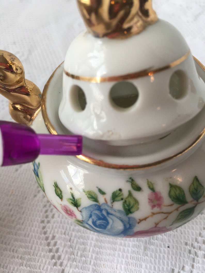 May include: A white porcelain incense burner with a gold trim and floral design. The lid has a gold bird finial and holes for the incense to burn through.