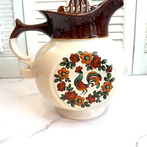 May include: A vintage ceramic pitcher with a brown lid and a rooster and floral design. The pitcher is white with a brown rim and a brown handle.