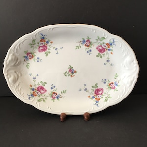 May include: White oval serving platter with a floral design featuring pink roses, blue and orange flowers, and green leaves. The platter has a gold rim and a slightly raised edge.