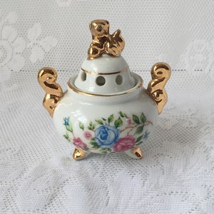 May include: A small white porcelain incense burner with a gold floral design and a gold cherub on top. The burner has a round body and three gold feet.