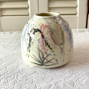 May include: A white ceramic vase with a round body and a wide opening. The vase is decorated with abstract black, blue, pink, and green brushstrokes and splatters.