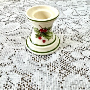 May include: A white ceramic candle holder with green trim and a holly berry design. The candle holder has a wide base and top, with a narrower center. The holly design features green leaves and red berries.
