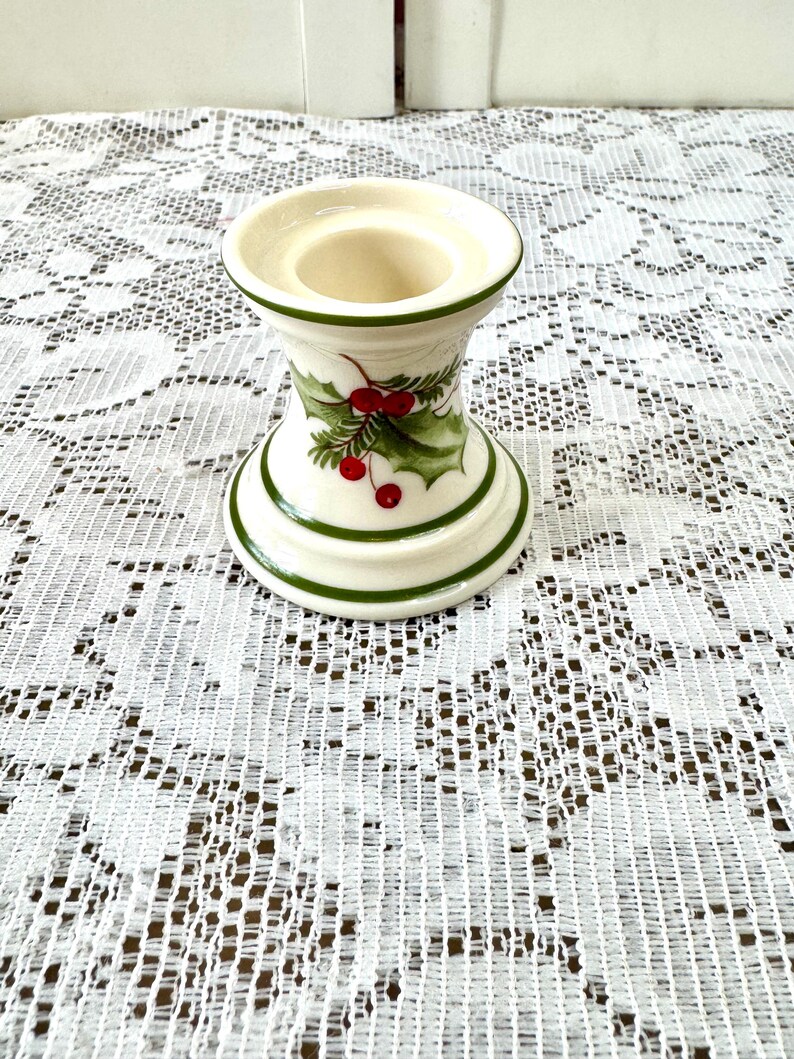 May include: A white ceramic candle holder with green trim and a holly berry design. The candle holder has a wide base and top, with a narrower center. The holly design features green leaves and red berries.