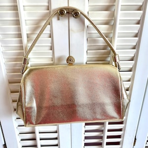 May include: A gold metallic evening bag with a jeweled clasp. The bag has a top handle and a rectangular shape.