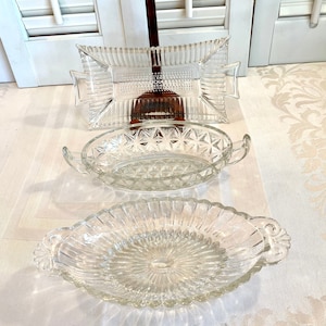 May include: Three clear glass serving dishes with different patterns. The dishes are oval shaped with handles on the sides. The dishes are on a white surface.