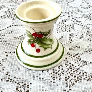 May include: Cream-colored ceramic candle holder with a green trim and holly berry design. The candle holder has a wide base and a flared top, perfect for holding a single candle. The holly design features green leaves and red berries.
