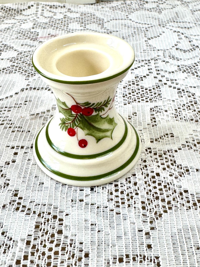 May include: Cream-colored ceramic candle holder with a green trim and holly berry design. The candle holder has a wide base and a flared top, perfect for holding a single candle. The holly design features green leaves and red berries.