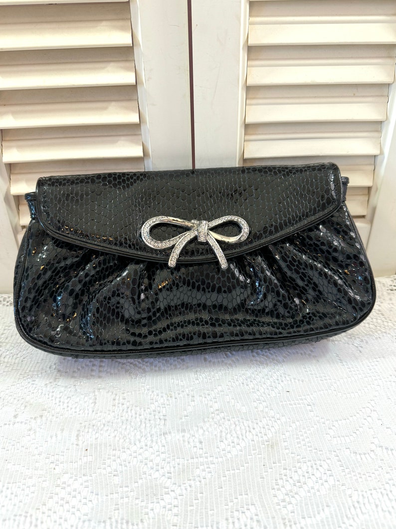 Op de afbeelding: Een zwarte clutch van slangenleer met een zilveren strik en strassversiering.