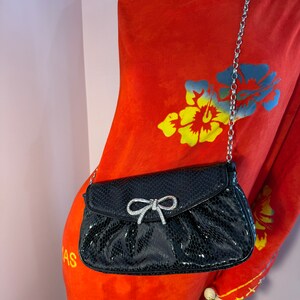 Op de afbeelding: Een zwarte clutch van imitatieleer met een zilveren kettingband en een detail van een strik met strass. De clutch is op een rode bloemenjurk.