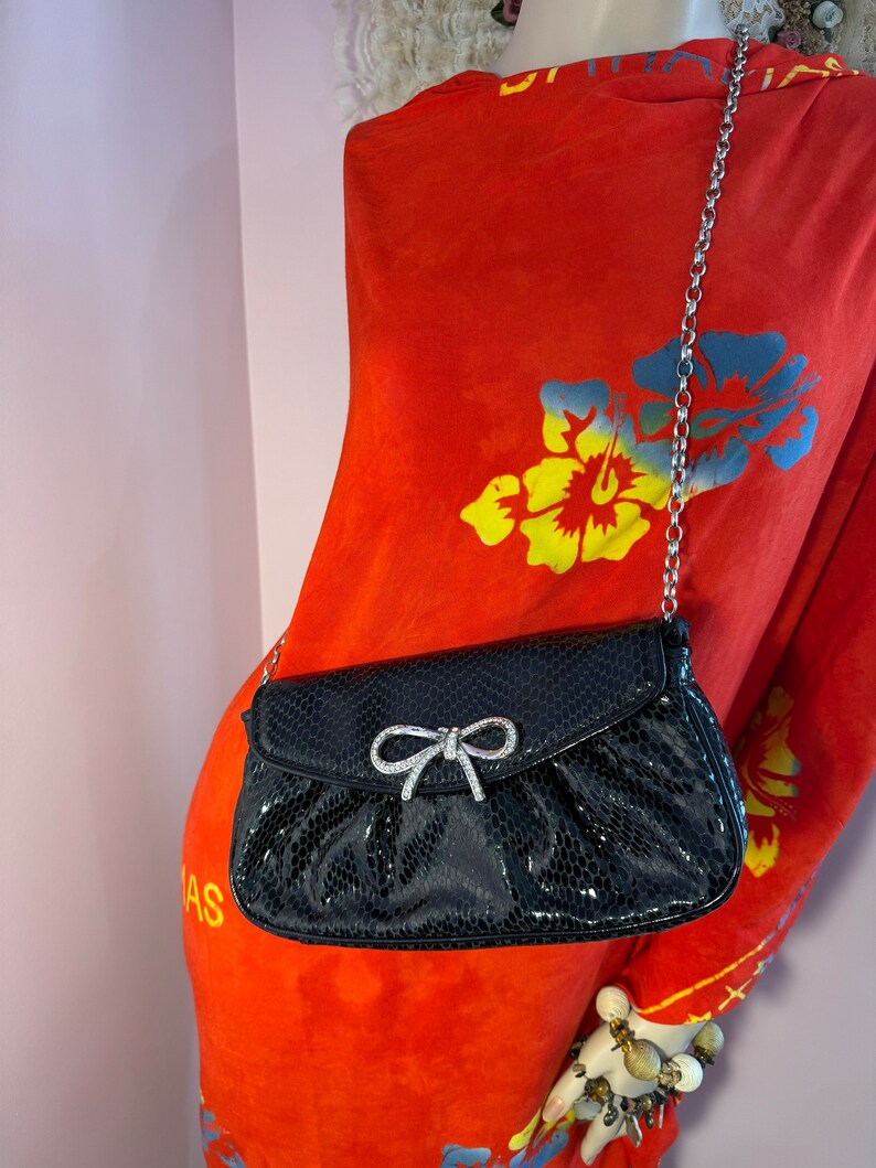 Op de afbeelding: Een zwarte clutch van imitatieleer met een zilveren kettingband en een detail van een strik met strass. De clutch is op een rode bloemenjurk.