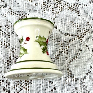 May include: A cream-colored, hourglass-shaped candle holder with green trim. The holder is decorated with holly leaves and red berries. The candle holder is a festive holiday decoration.