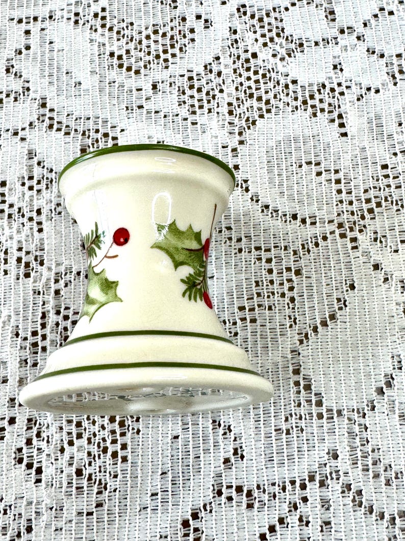 May include: A cream-colored, hourglass-shaped candle holder with green trim. The holder is decorated with holly leaves and red berries. The candle holder is a festive holiday decoration.