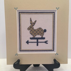 May include: A framed cross-stitch of a brown rabbit on a weathervane with arrows pointing in opposite directions. The frame is beige with a blue and white border. The background is a light pink.
