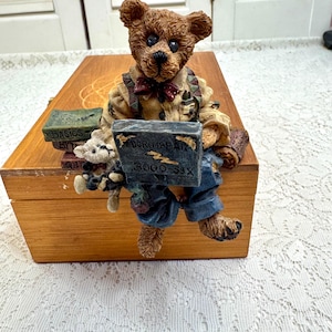 May include: A brown teddy bear figurine wearing a shirt, overalls, and a bow tie, holding a book that reads "MICRO-BEAR 3000-SIX". The bear is sitting on a wooden box with books that read "BASICS BYT".
