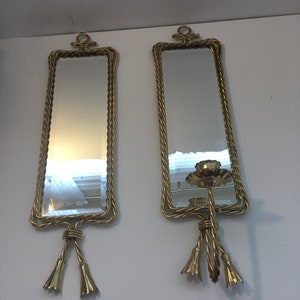 May include: A pair of rectangular wall mirrors with gold-toned metal frames featuring a twisted rope design. Each mirror has a decorative top loop and a bottom section with two dangling bell-shaped accents. One mirror includes a candle holder.