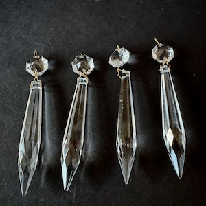 May include: Four clear crystal chandelier pendants with faceted edges and gold-colored metal accents. Each pendant has a multifaceted top and a long, pointed bottom. The pendants are arranged against a dark background.