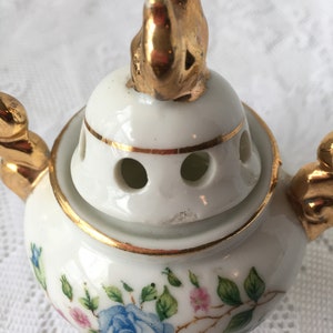 May include: White porcelain incense burner with a gold trim and floral design. The lid features a gold figurine of a sitting animal.