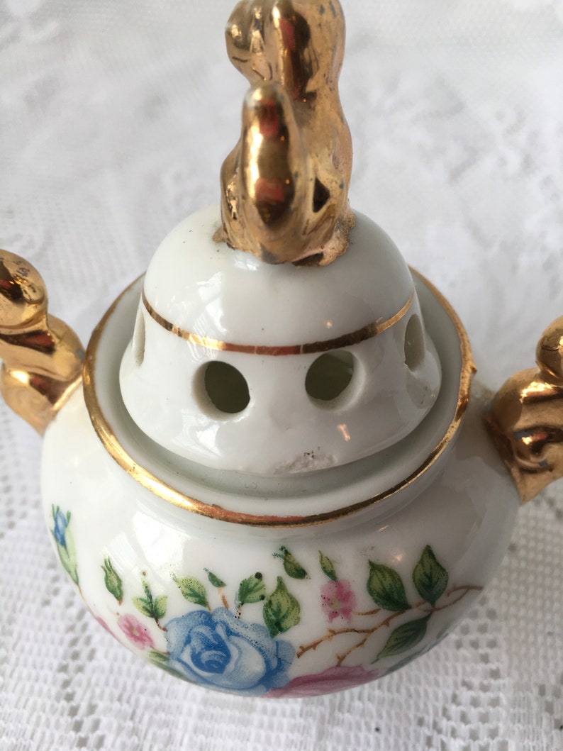 May include: White porcelain incense burner with a gold trim and floral design. The lid features a gold figurine of a sitting animal.