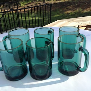 May include: Six teal green glass mugs with handles. The mugs are on a white surface.