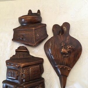 May include: Three vintage wooden wall decor pieces in the shape of a stove, a bellows, and a coffee grinder. The pieces are painted brown and have a rustic look.